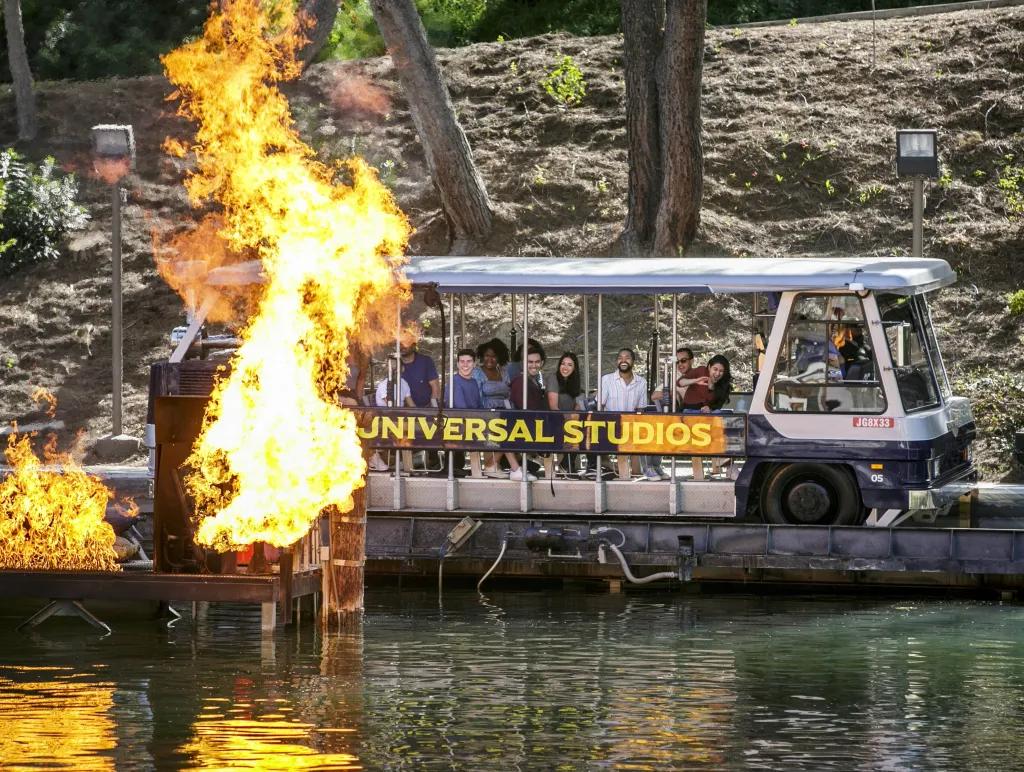 Lake with a large car with rows of people on, on the side of this vehicle it says 'Universal Studios'. Over the water there is a wooden pier which is exploded on fire.