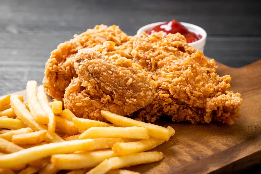 Crispy piece of fried chicken and fries on a wooden board next there is a white circular pot of ketchup.