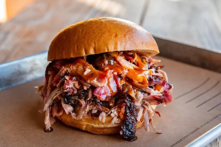 Large burger full of puller pork, bacon and a red sauce served in metal tray.