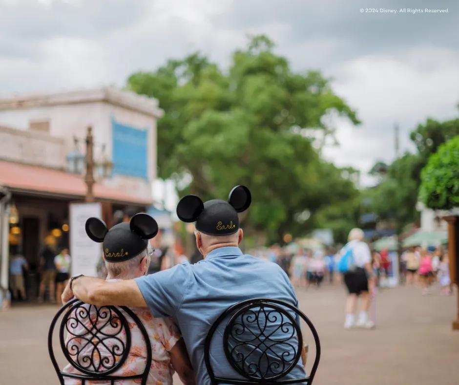 A picture from behind a couple sat on black garden chairs in the Animal Kingdom Theme Park, both wearing mickey hats with their names personalised onto them. The man has his arm around the woman and they are looking towards the Tree of Life.