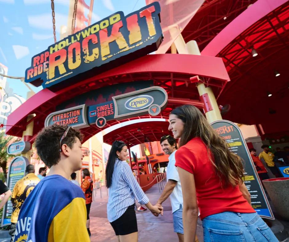 Guests entering the express queue of a red and yellow coloured coaster, a couple with their two teenage children. Above their head is a sign saying 'Hollywood Rip Ride Rockit'.