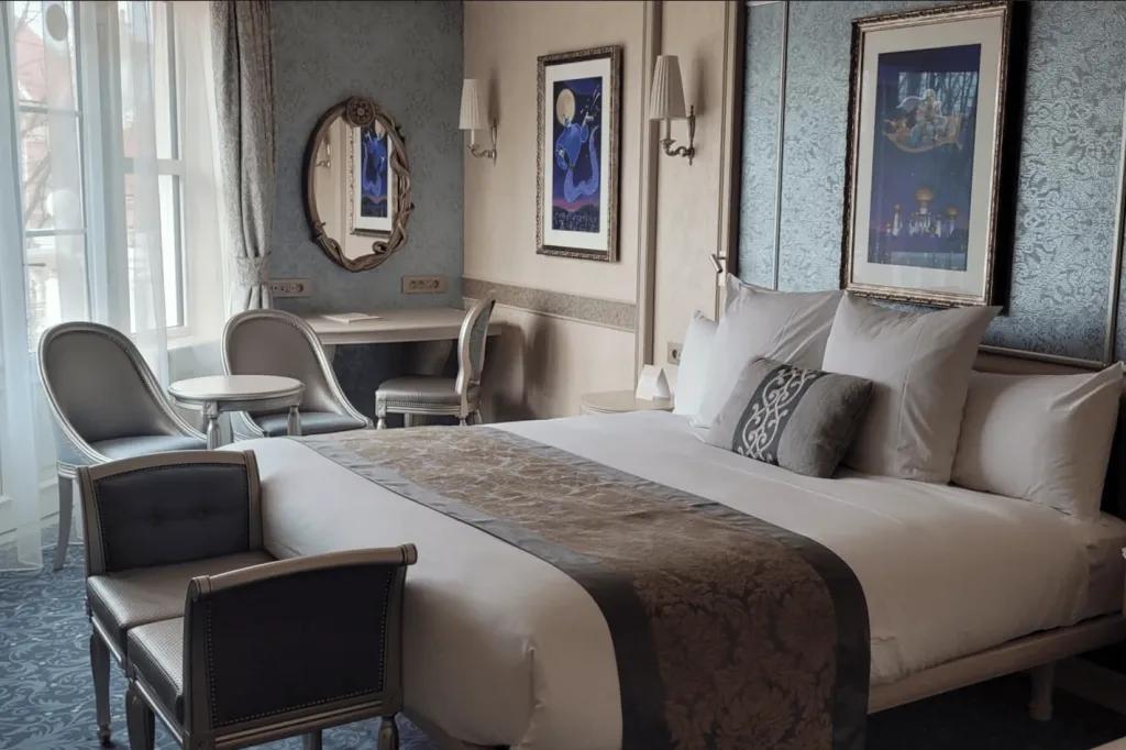 A hotel room with a king-sized bed in the centre next to a balcony door and a table and chairs