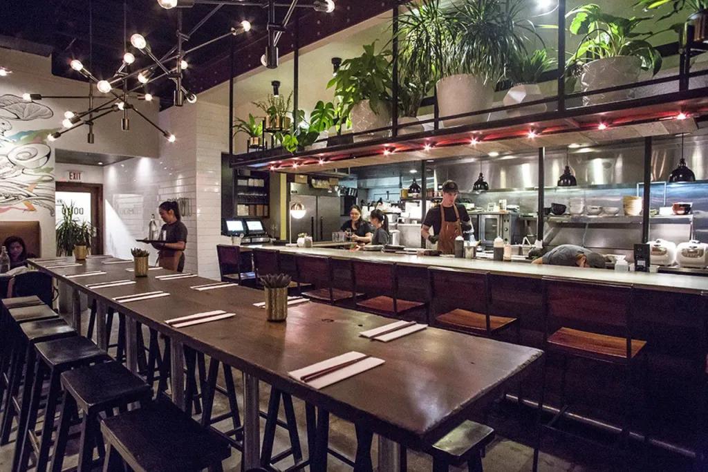 Long wooden table with black stool chairs in modern restaurant, with a waitress stood in the background and a clear into a kitchen, where a chef is stood.