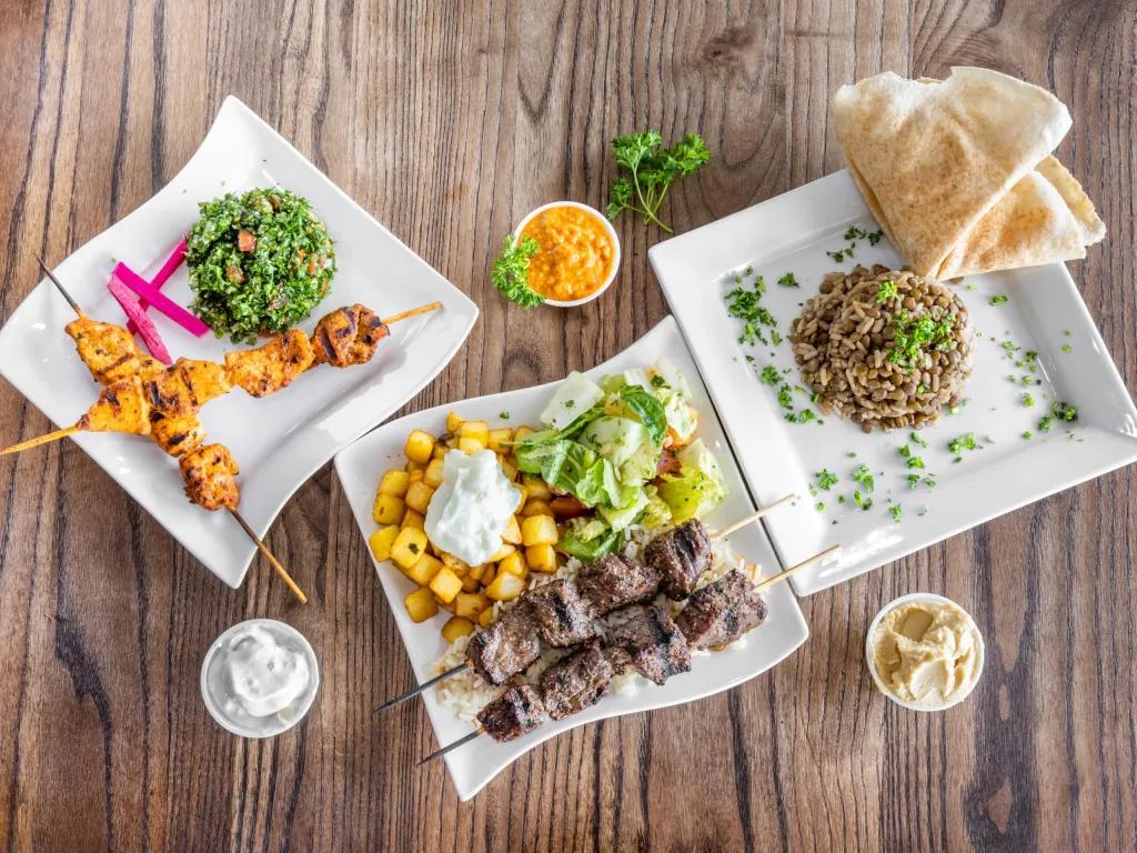 Three square plates on a wooden table, one with chicken kebabs with vegetables, another with lamb kebab and potato chopped into small squares and a salad and last of all, a plate with brown rice mixed with beans. There are three small pots of sauces.