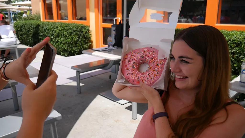 Someone taking a photo of a woman smiling holding a giant doughnut with pink icing and sprinkles on it. The doughnut is in a white paper box which she is holding up to the right of her head.