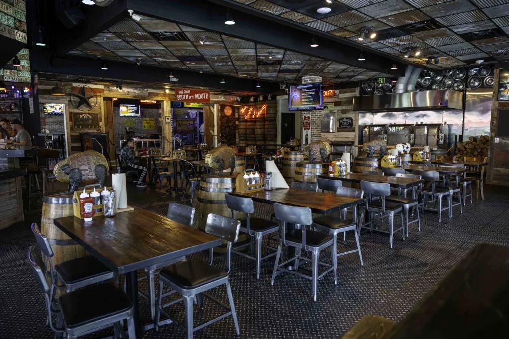 Darkly lit restaurant with black metal chairs surrounding dark wooden square tables with southern American country theming. There are barrels and neon signs on the wall with a tv playing sports events. 