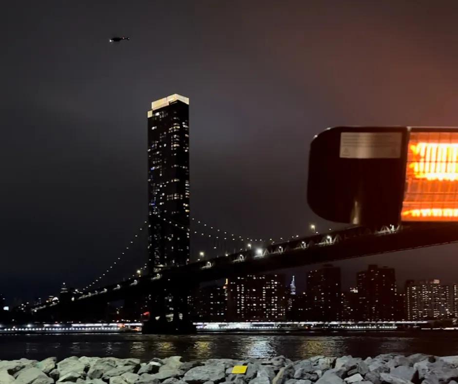 To the right there is a heater and to the left there is an incredible view from a restaurant with skyscrapers and the Brooklyn Bridge lighting the night sky. There is also the river below and above a helicopter is flying.