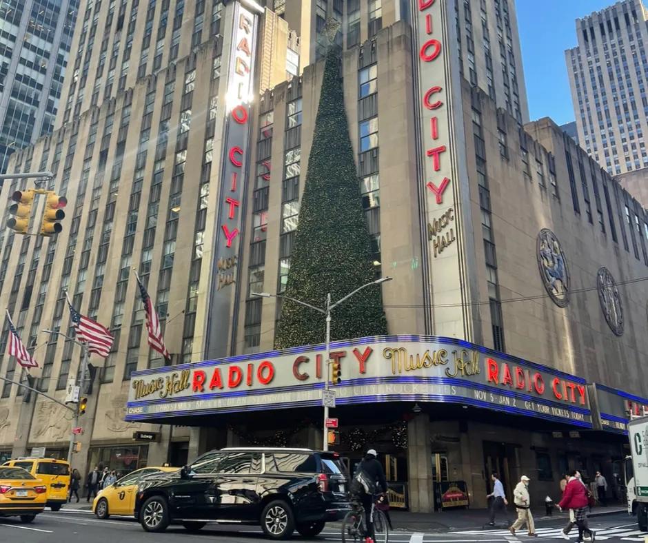 Cars driving along the road below building with silver, red and blue sign saying "Radio City Music Hall'. There is a giant half christmas tree on top of the sign leaning against the wall. On the right there are American flags waving in the wind.