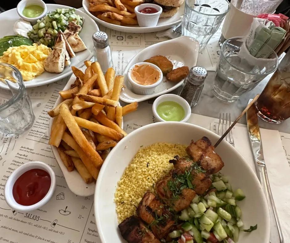 Table with a lot of food on it. There are white bowls with chips, fried pickles, chicken kebab, salad and eggs with avocado.