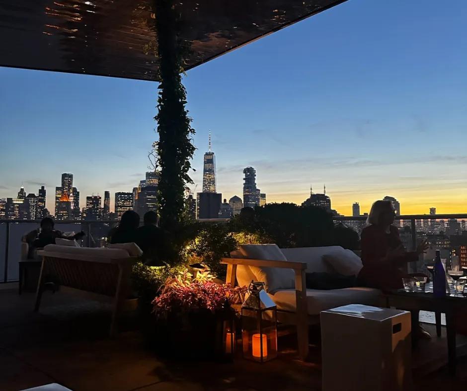 Terrace with many plants and flowers on post holding up roof, in the background there is a sunset over the city, with buildings lit up. There are people talking whilst sat on clean cream sofas.