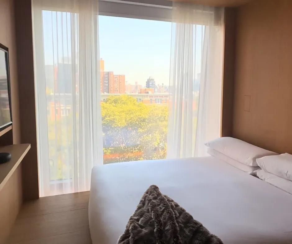 Bed on wooden floor with white sheets next to a large window overlooking New York City. There is a television on the wall in front of the bed and white curtains slightly drawn closed.