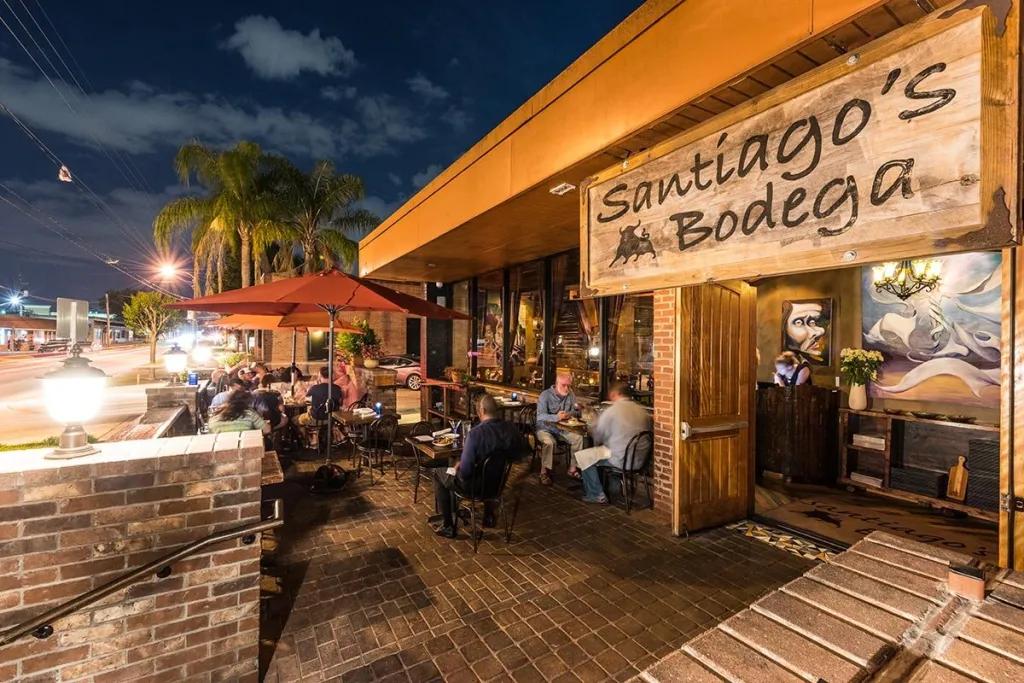 Outside of a restroom with grey brick terrace and people sat beneath red umbrellas. There is a wooden sign which says Santiago's Bodega in black.