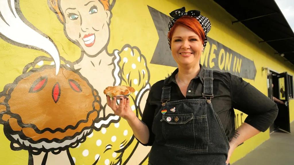 Woman with red hair stood holding pie in front of building with yellow and black art piece painted on side of woman holding a steaming hot pie.