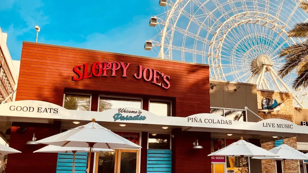 Box shaped restaurant coloured red and beige with a lighter red sign reading "Sloppy Joe's" with the white Orlando Eye in the background.