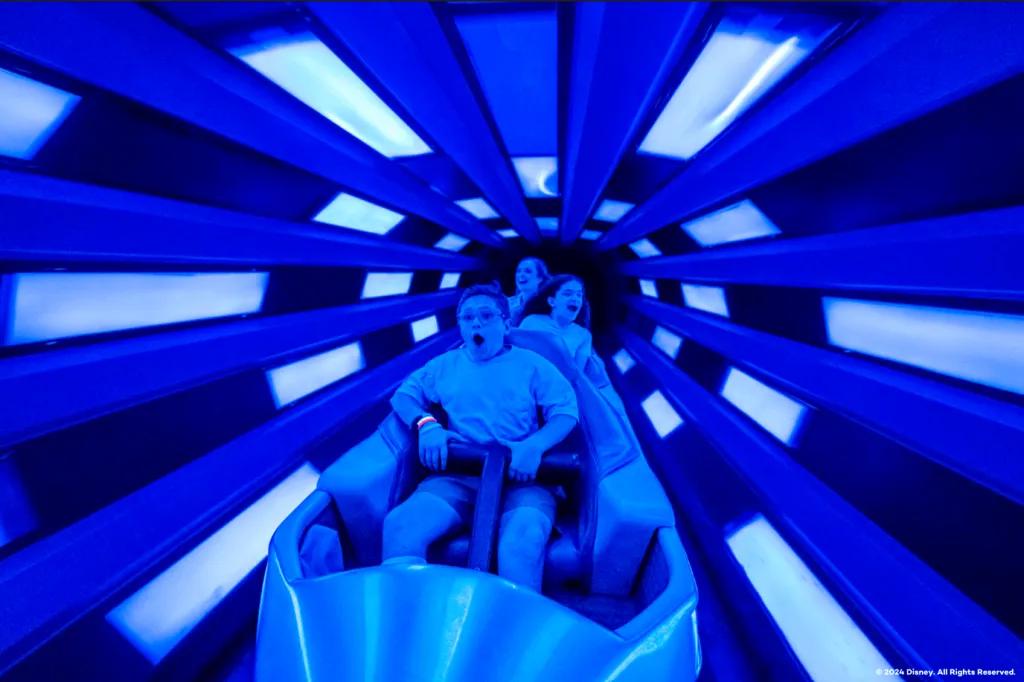 Three people sat one in front of the other on a rollercoaster vehicle in a tunnel lit blue