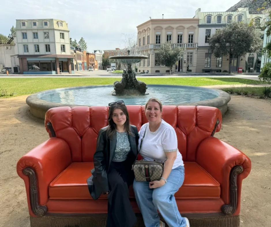 A young woman in blue sparkly top sat next to woman in white t-shirt sat on orange sofa in front of the iconic circular fountain from Friends credits.