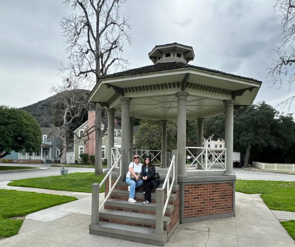 Gazebo in the centre of a town with while pillars, a brick staircase and grey outlines. In the background there are tall trees without leaves on them, a mint green and white house and a blue house with white fences.