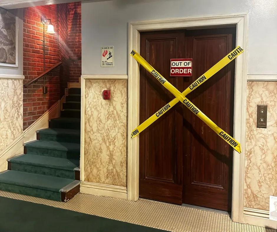 Apartment staircase with brick walls, dark blue staircase and a beige and white wallpaper. There is a lift with a dark wooden door, an 'OUT OF ORDER' sign on the doors and two strips of caution tape in an X shape on the door.