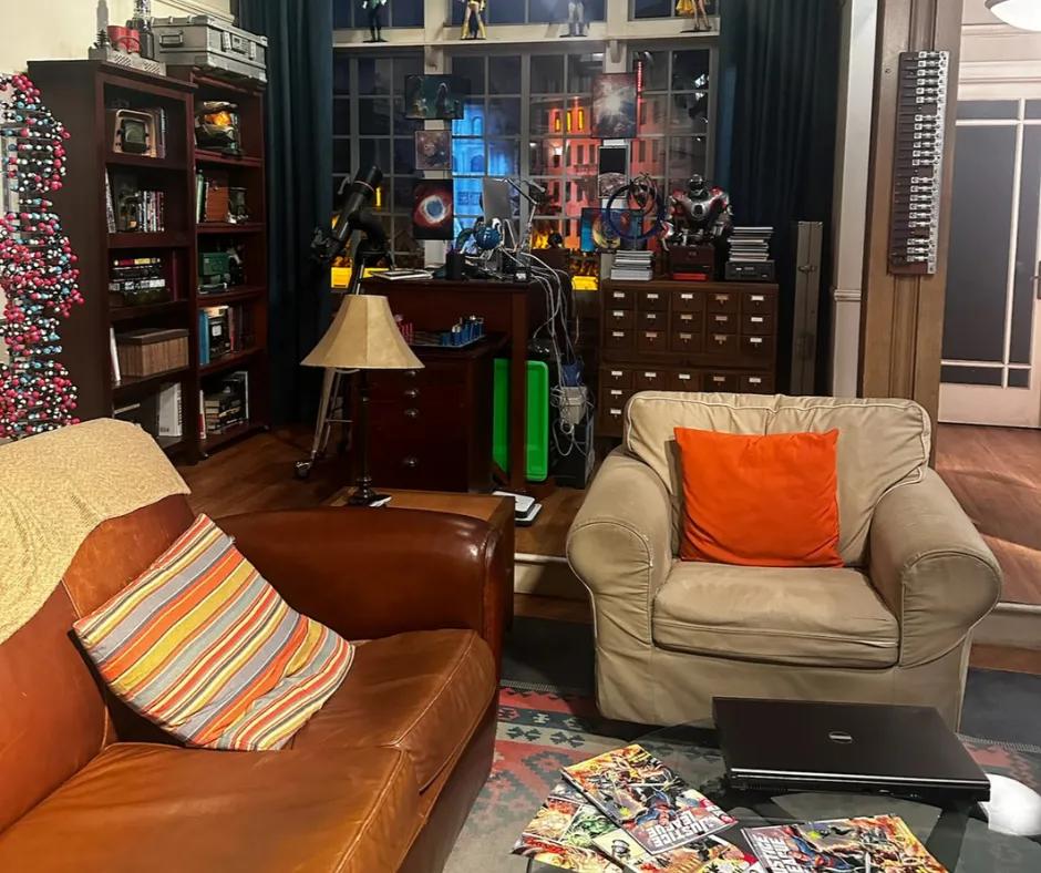 Apartment designed set, with dark brown sofa with patterned blanket on top and stripey cushion on. Another smaller sofa which is grey with an orange cushion. In the background there are DNA structure statues, comic books spread out on coffee table and robots on dark wooden drawers. To the right there is a white door with glass.