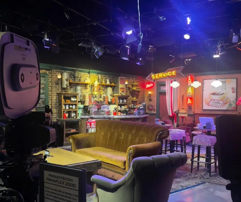 Set of the Friends coffee shop, to the left there is a film camera with the iconic coffee table and brown sofa behind. There is also the coffee counter with cake holders and signs saying 'service' hanging up. Behind there are other tables and some stools.