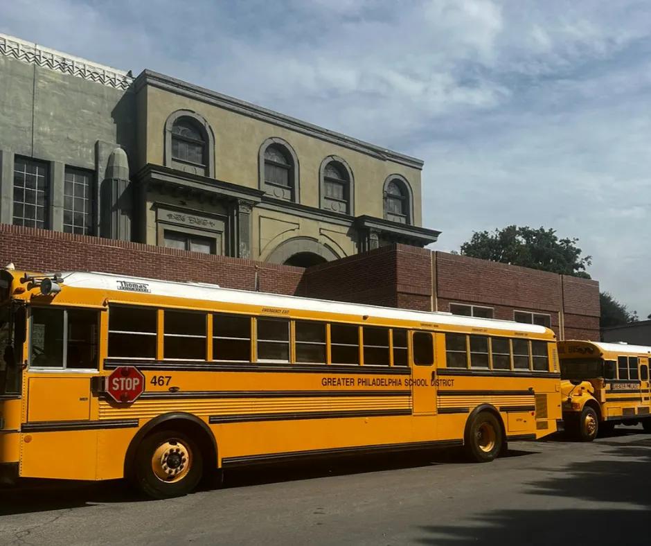 Two large yellow school buses with a stop sign attached to one and writing along in black saying "GREATER PHILADELPHIA SCHOOL DISTRICT". In the background is a brick building and behind that is a couple more buildings.