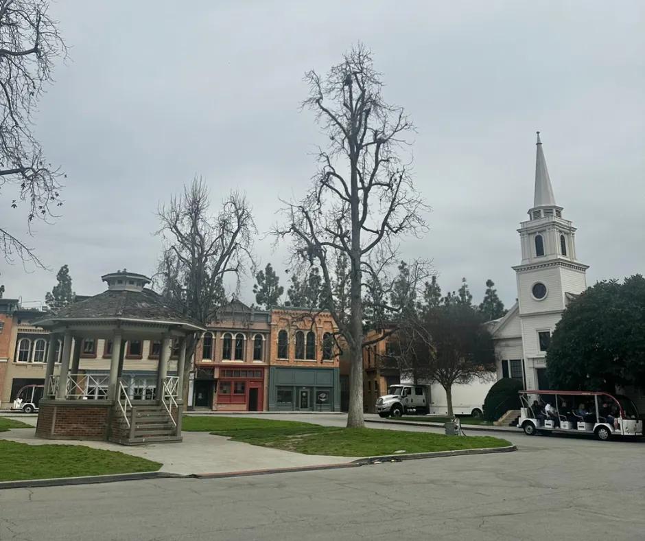 White gazebo to the left, with buildings of varying colours behind such as red and dark blue store fronts below. A large white lorry is next to the white church with pointed roof and circular windows. 