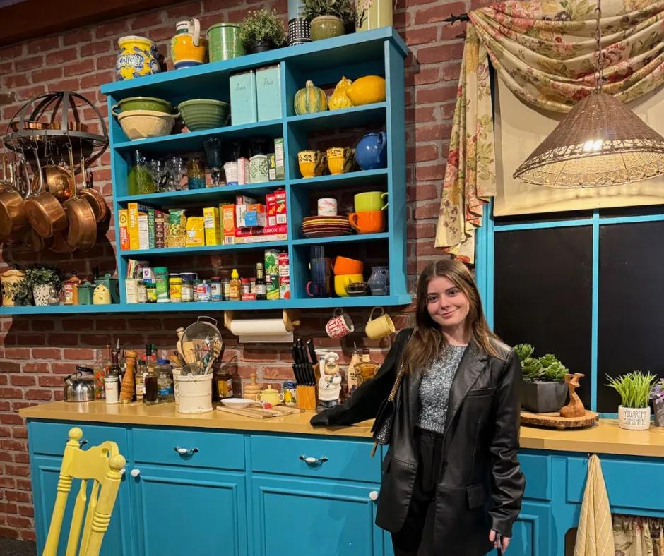 Woman in sparkly blue top and black blazer stood with hand on wooden kitchen counter that has light blue draws. Above there is cluttered utensils, condiments, pans hanging up and shelves with cutlery on.