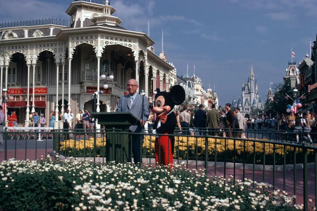 Old image of main street and Cinderella's Castle and in front Mickey is stood with Walt Disney as he delivers a speech.