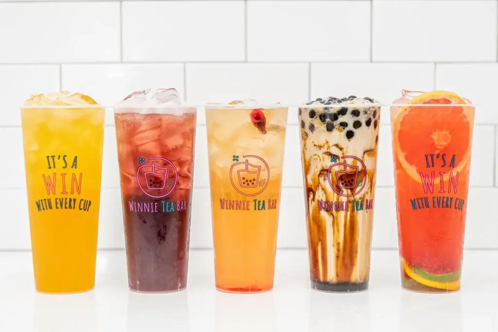 Five large clear cups of different drinks, from left to right, there is a yellow drink with ice saying "It's a win with every cup", a dark red drink with ice with the logo on it, an orange drink with ice, a brown milk tea with small black tapioca pearls in and lastly a pink drink with slice of orange in.