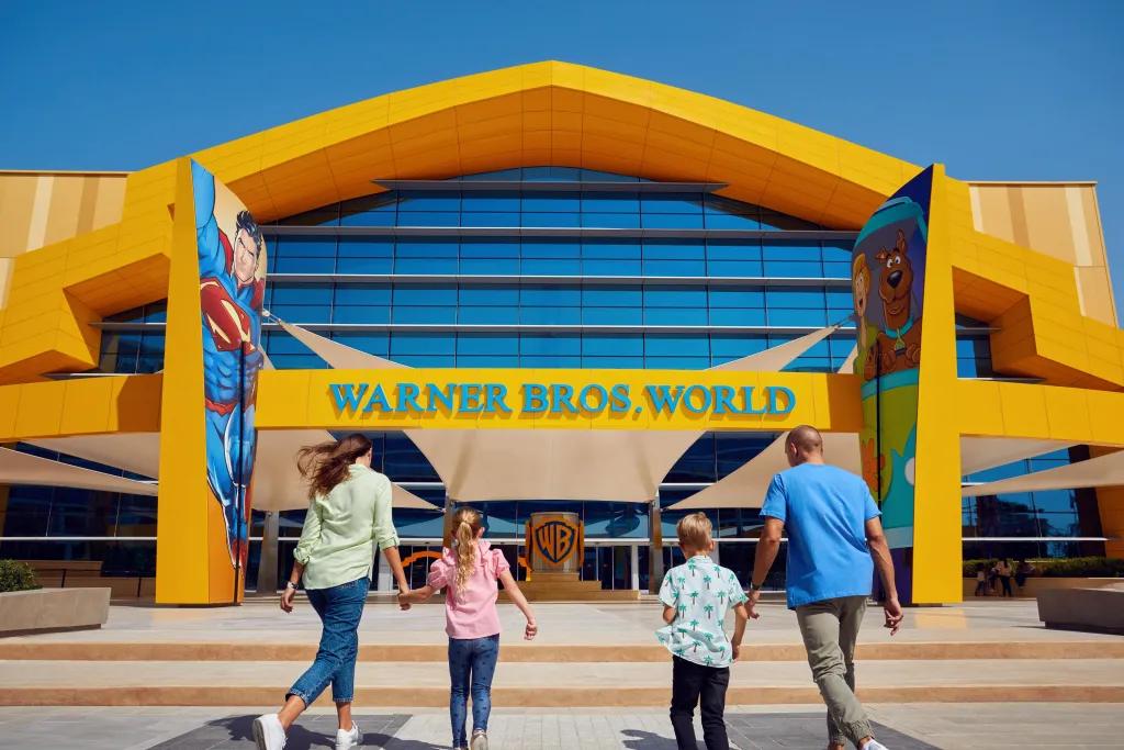 Family of four walking towards a large yellow building with two images either side of the entrance, one of superman comics and one of scooby doo. There is a blue sign saying "Warner Bros. World" and below there is a copper WB sign.