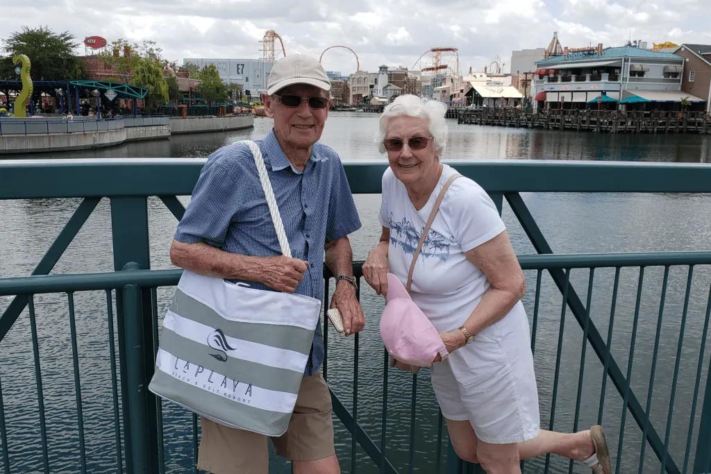An older couple posing in front of a lake with a theme park in the background