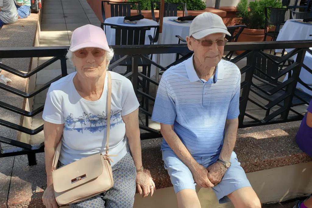 An older couple smiling at the camera while they sit on a low wall