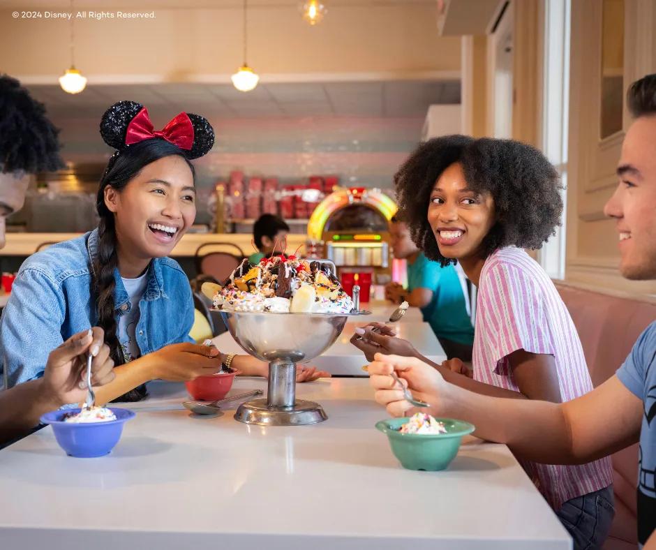 4 guests sat around a huge silver bowl full of many scoops of ice cream, whipped cream, sprinkles, m&m's brownies and cherries. One girl has sparkly minnie mouse ears on her head and both women are looking at one of the men. They are all holding spoons near their multicoloured mini bowls.