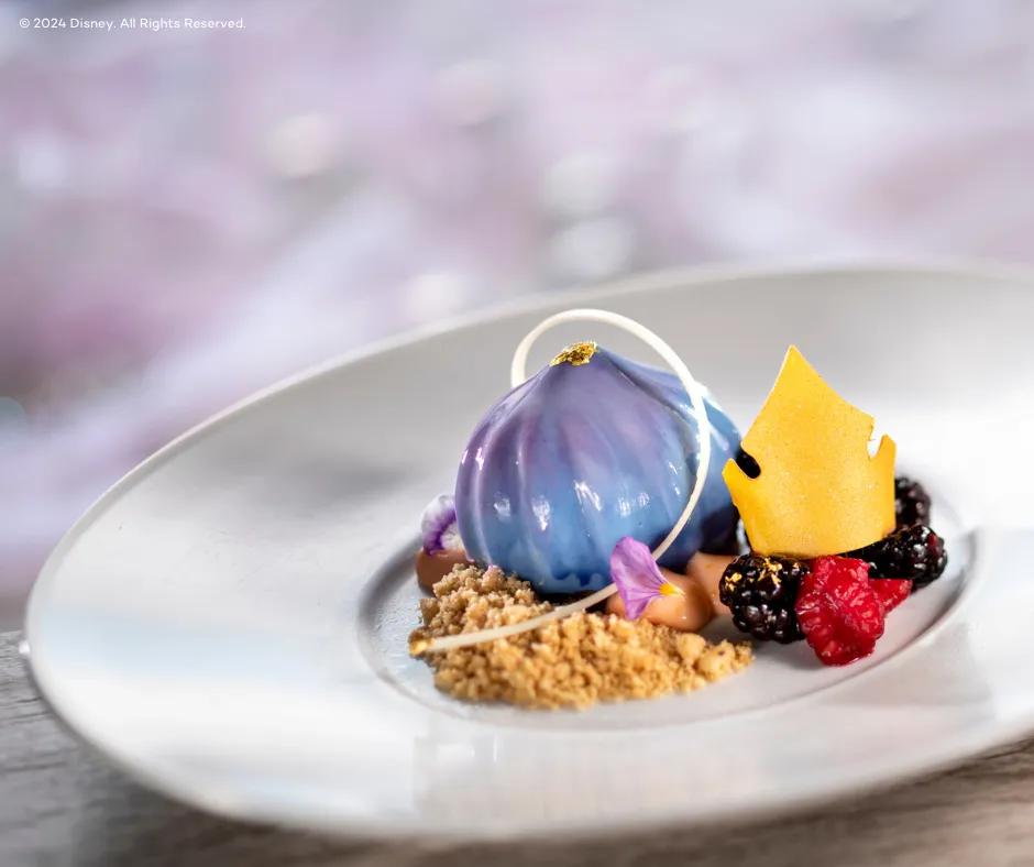 A white plate of gorgeous food, what looks like a cheesecake from a fairy tale. There is a shiny blue and purple coloured tear shaped food on top of small crushed pieces of buscuit crumbs. There is a white chocolate arch over this and to the right there is a yellow crown shaped edible paper on top of raspberries and blackberries with small pieces of gold on them.