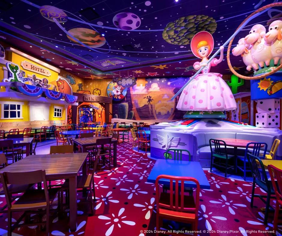 Empty restaurant with large statues of toys including bo peep and her three sheep, Bo peep is a blonde doll wearing a white dress with pink details. There are also large dice statues. The tables are wooden and multicoloured in Toy Story colours. The backdrop is fake paper town based on Woody's backstory and the ceiling has space theming.