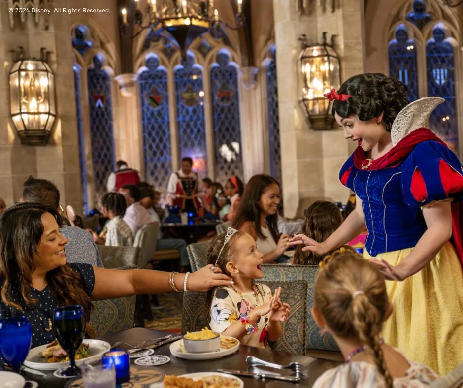 Princess dressed in blue and yellow dress with red details on it and a red cape as well as red bow in her black hair (Snow White) greeting guests at a table in a restaurant. A little girl looks up to the princess smiling and shocked with a tiara in her hair as her mother amiles watching, pulling back a strand of hair from the child's face. There is also an older sister looking at the princess with a plait in her hair.