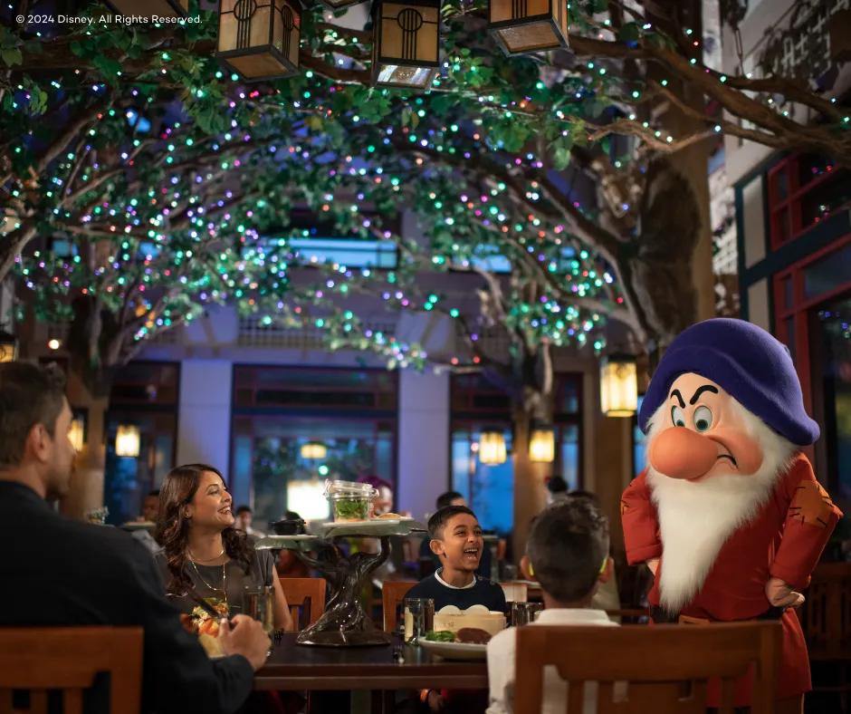 A restaurant with dark lighting yet cosy, above there are decorative trees inside with multicoloured twinkling lights, below there is Grumpy the dwarf, a man with a large nose and furrowed eyebrows wearing a blue hat and a red outfit. Grumpy has his hands on his hips and is looking towards guests sat at a wooden table, a family of 4, a mum, dad and two boys who are smiling/laughing at him.