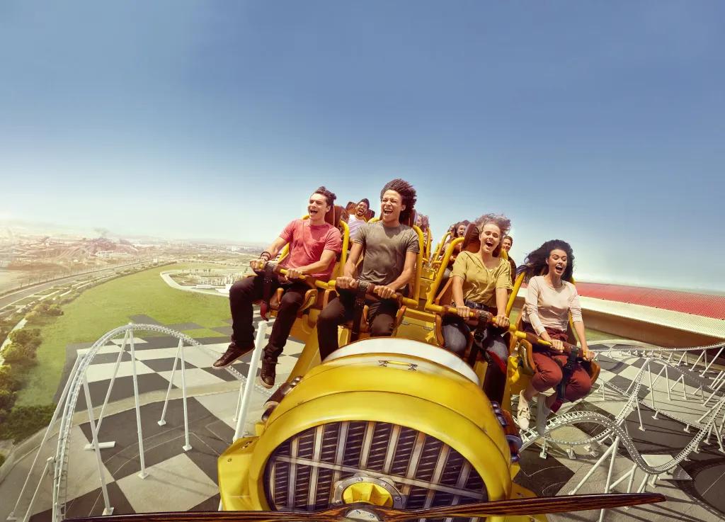 White rollercoaster track on black and white floor with yellow coaster car and four people screaming and smiling whilst clinging onto the lap bars.