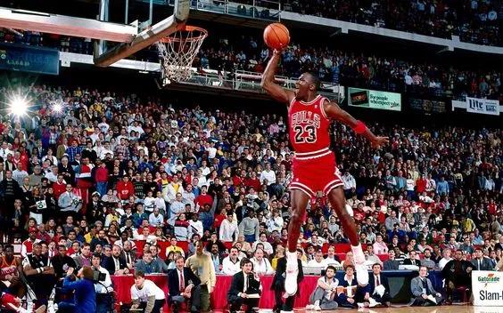 Man in number 23 red basketball uniform floating mid air about to dunk the basketball into the net elevated high above the ground. In the background you can see the audience engrossed watching.