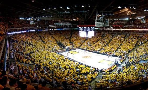 A white rectangular court with a large 4 sided tv screen hanging above and a full audience sat in tiered seating around. All audience members are wearing yellow t-shirts.