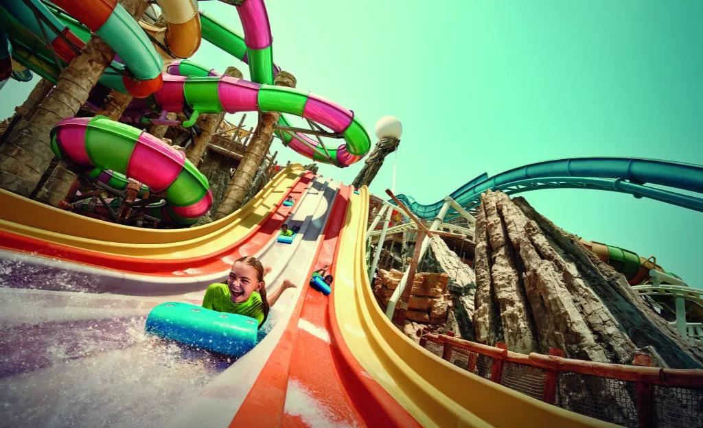 A huge pink and green stripey waterslide to the left, a large steep blue waterslide to the right and down the centre there is a racing waterslide where kids lay on stomachs on mats down small hills. There is a little girl winning, smiling with joy.