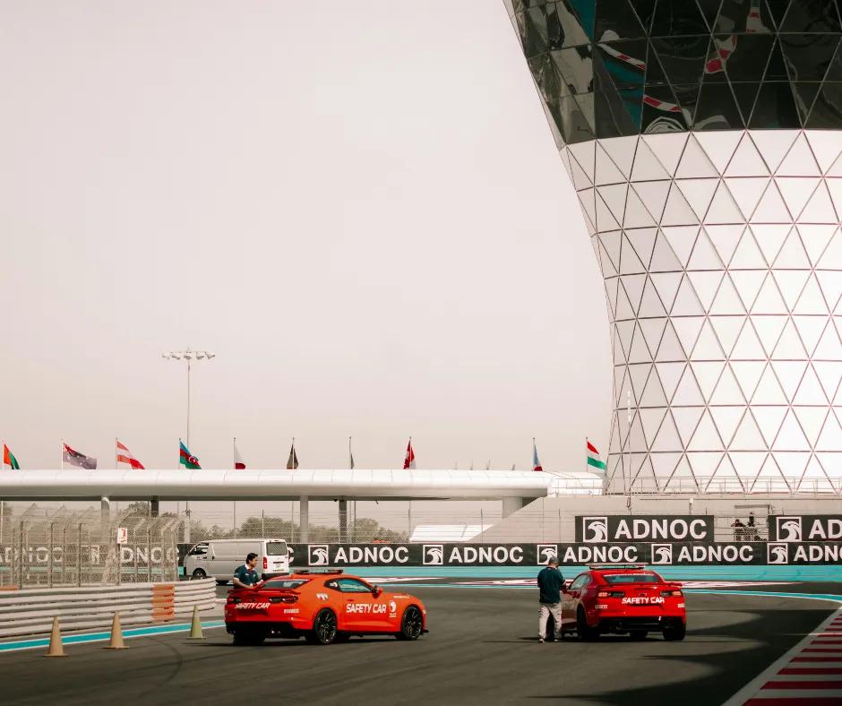 Two red cars on race track with two men stood next to either one. There is a white structure in the background with black windows and then flags lining the track.