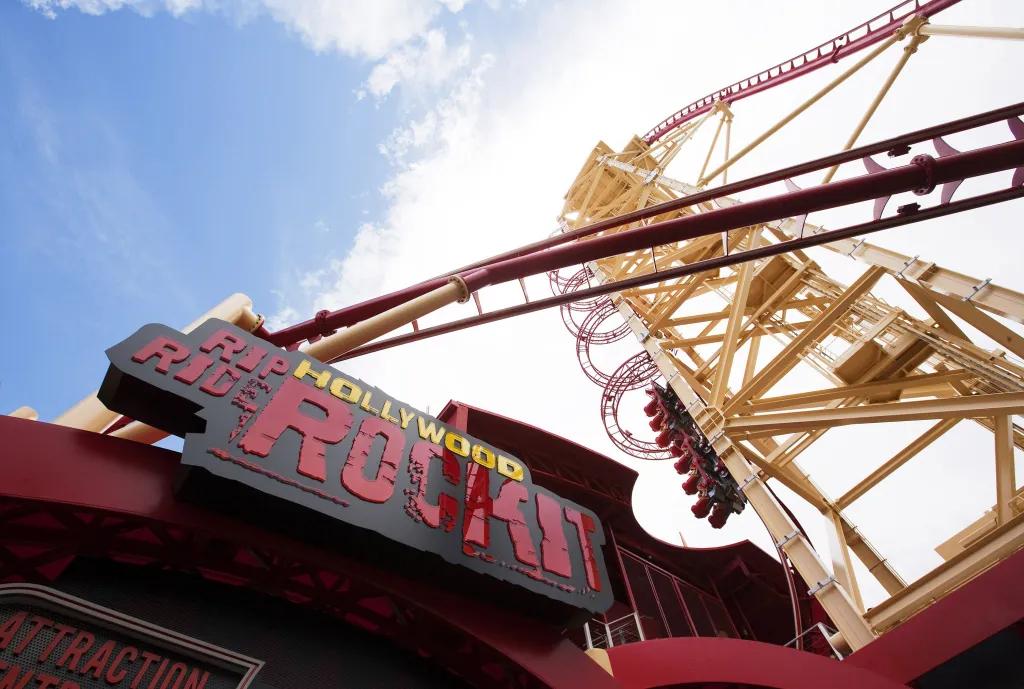 Rollercoaster red cart heading up a vertical chain lift surrounded by circular steel arches, almost about to drop down the steep red and yellow track. There is a black sign with red and yellow text saying "Hollywood Rip Ride Rockit"