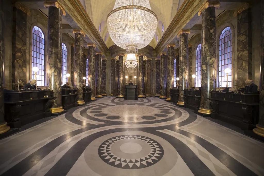 Large clear building with black and white tiled patterned floor. Above hangs two grand chandeliers. Along the left and right hand side are tall dark brown desks with 3 goblin characters sat at them working. In the middle there is a larger desk with the main goblin working. This is a room themed as an elaborate bank. 