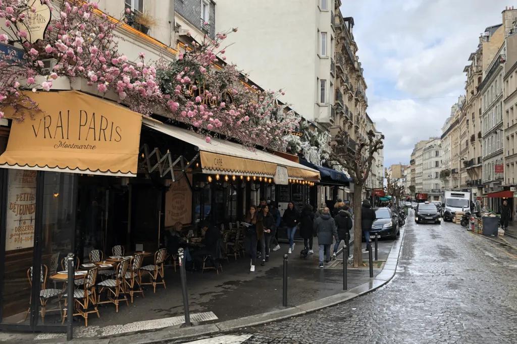 A wet road in Paris lined with parked cars and the outside of s restaurant which is named "Vrai Paris". Above the beige cover there are pink flowers and below there are tables with chairs.