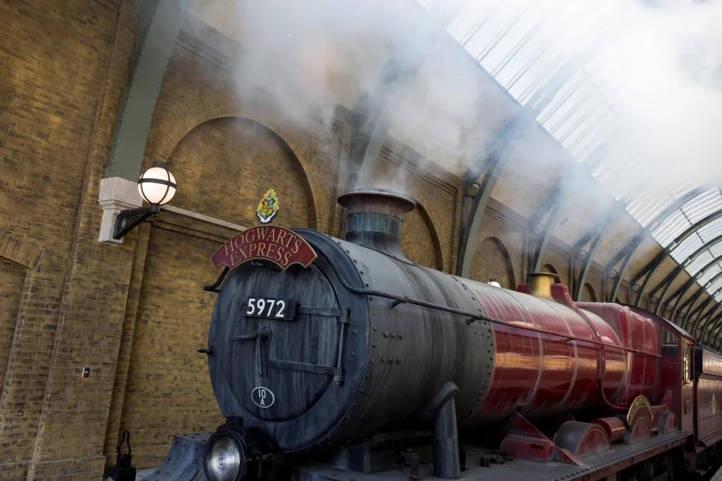 Red and black train with steam coming out of its funnel is still in a brick station. On the front it says "Hogwarts Express"
