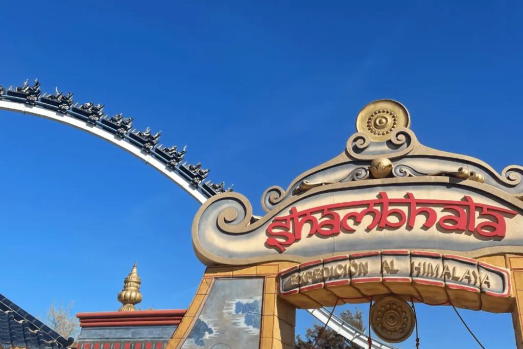 A large sign which is shaped as an archway which says shambhala - expedicion al Himalaya. The sign is coloured red and gold with patterns and artistic drawings of people on it. Behind there is a large white rollercoaster with a black rollercoaster carriage on it.