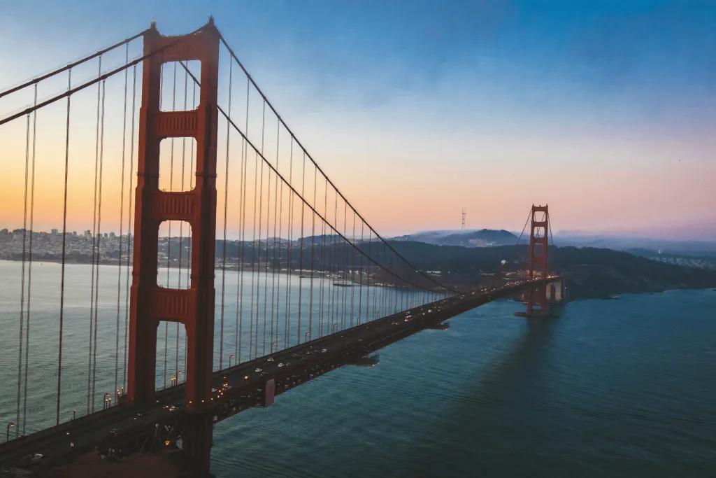 The sun is setting with blue and orange hues. Over a large river, there is a large red suspension bridge