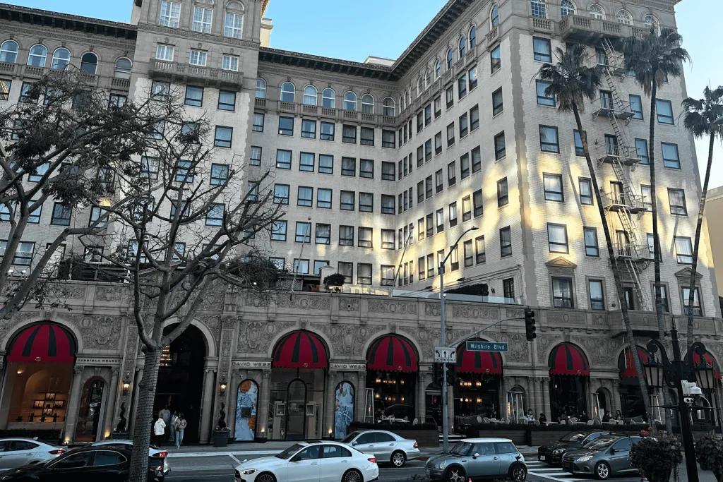 A large clean building on the side of a busy road with palm trees in front of it. At the front of this building there are covers over windows coloured red and black.
