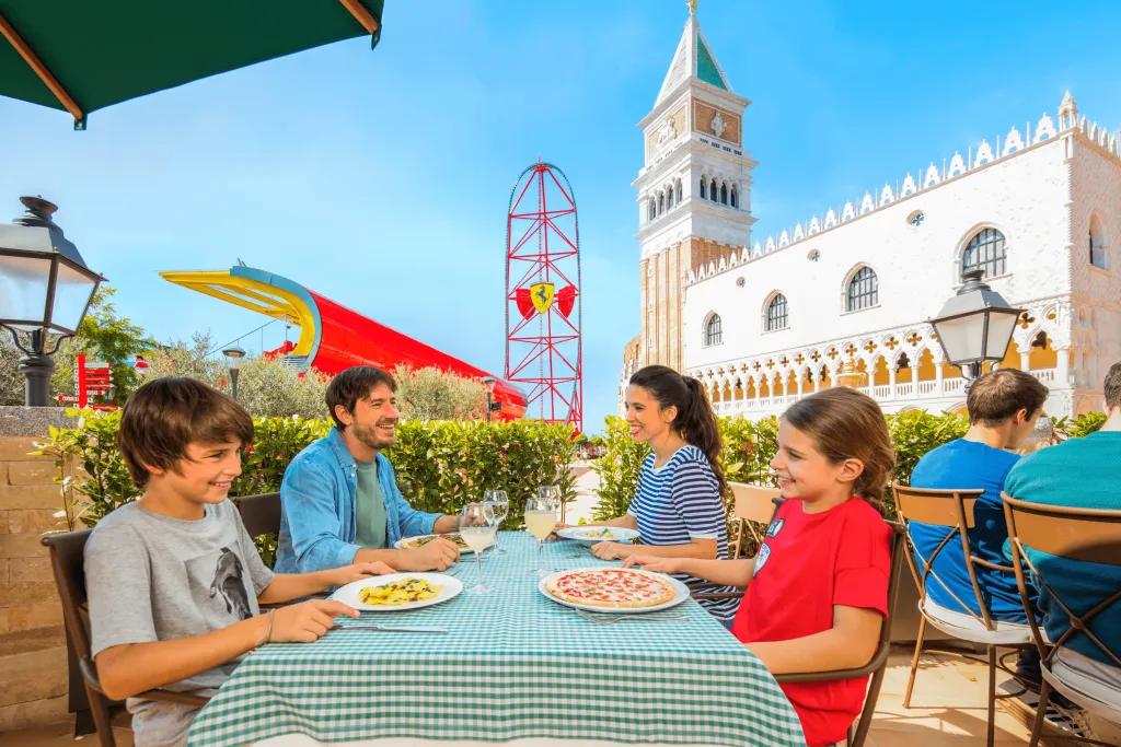 A family of four, a dad, mum, son and daughter, all sat at table with green and white checkered table sheet eating pizza and pasta with a beige, white and green building behind them. To the left of the building there is a tall red rollercoaster with a Ferrari sign on the middle (yellow shield shape with black horse)